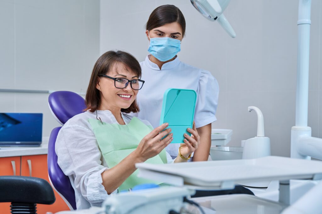 Happy middle aged woman together with dentist, patient sitting in dental chair looking at mirror. Prosthetics, treatment, implantation, dental teeth health, beauty care concept