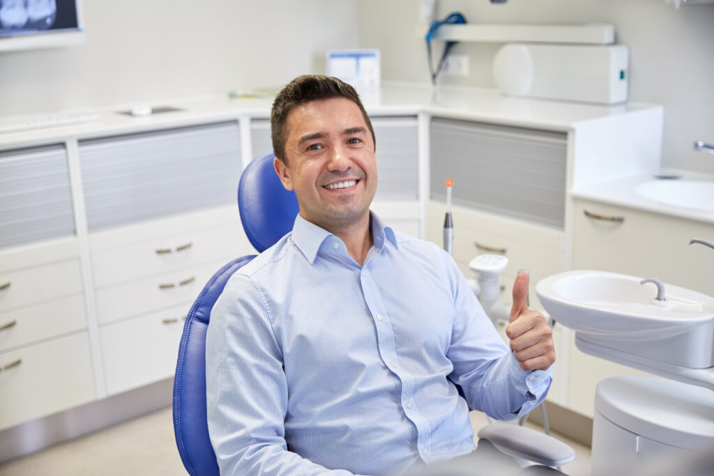 people, medicine, stomatology and health care concept - happy male patient sitting on dental chair and showing thumbs up at clinic office