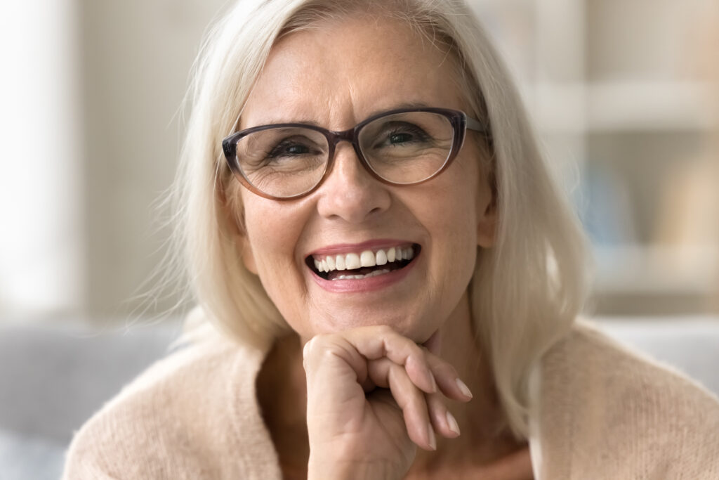 Face of happy senior female model with stylish elegant glasses, toothy smile, fair hair. Cheerful old woman looking at camera for close up portrait, laughing, showing healthy white teeth