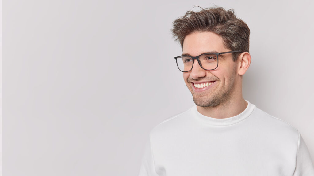 Happy man has bristle and dark hair looks gladfully away wears transparent glasses and casual jumper isolated over white background with copy space for your promotional content. Emotions concept
